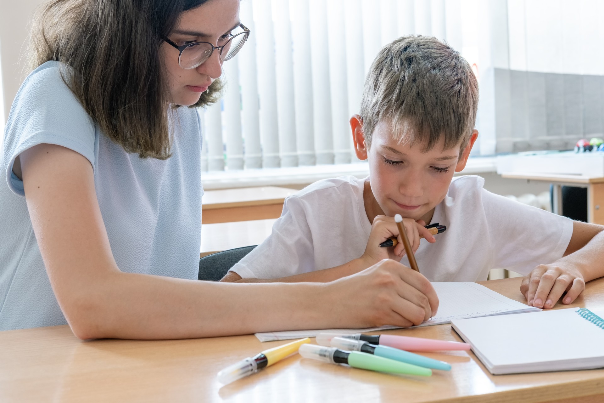 A boy and a teacher doing homework, writing text in a notebook at the table. A mother helps her son write in a notebook